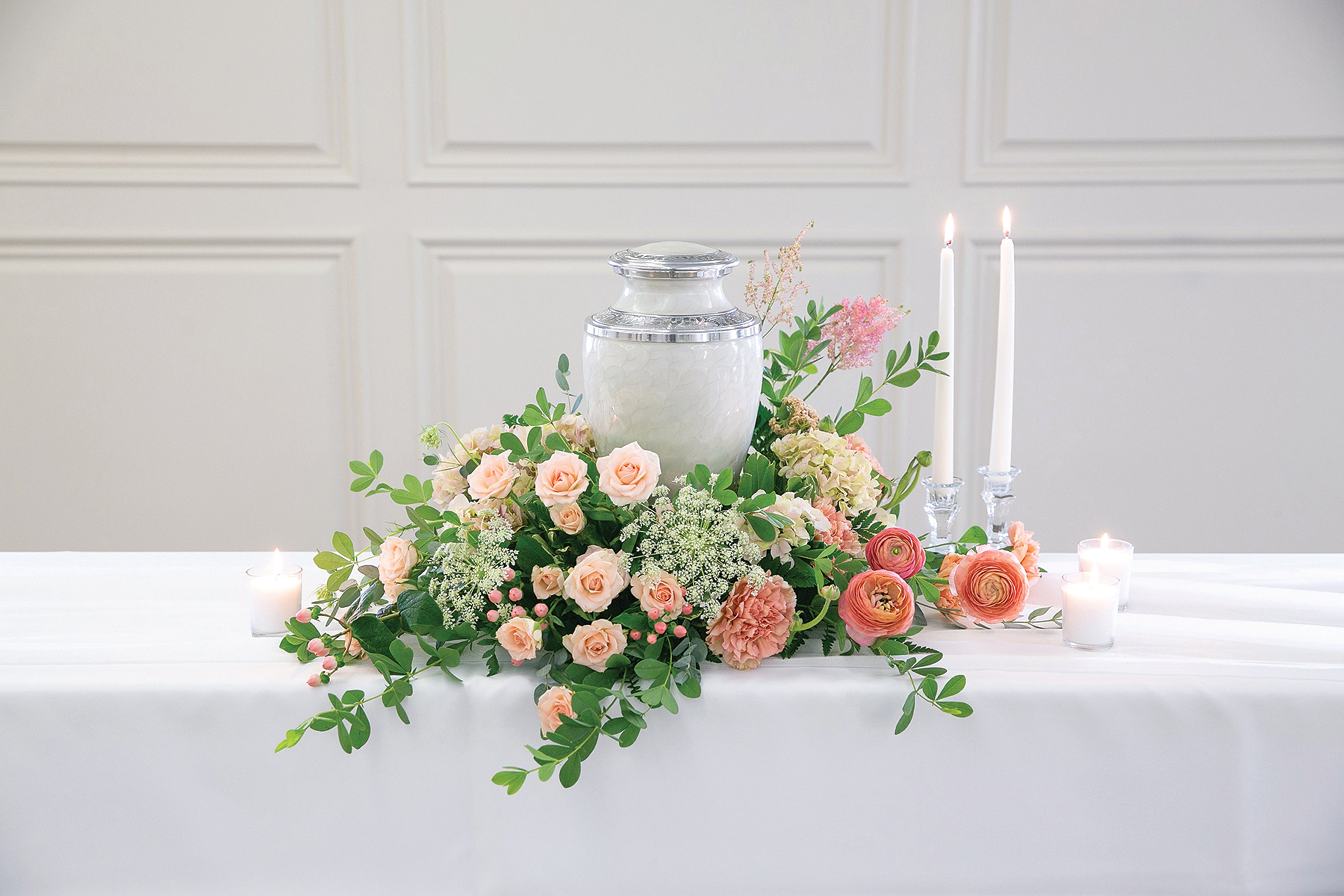 A floral centerpiece arrangement with a white urn, peach and pink roses, green foliage, white candles, and small white candles on a white table against a white paneled wall.