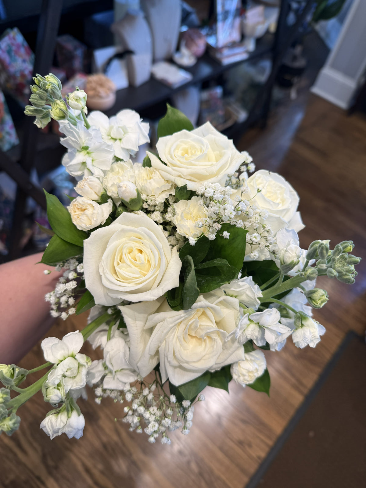 A bouquet of white roses and white baby's breath flowers with green foliage, held by a person's hand in a home setting with a wooden floor and furniture in the background.