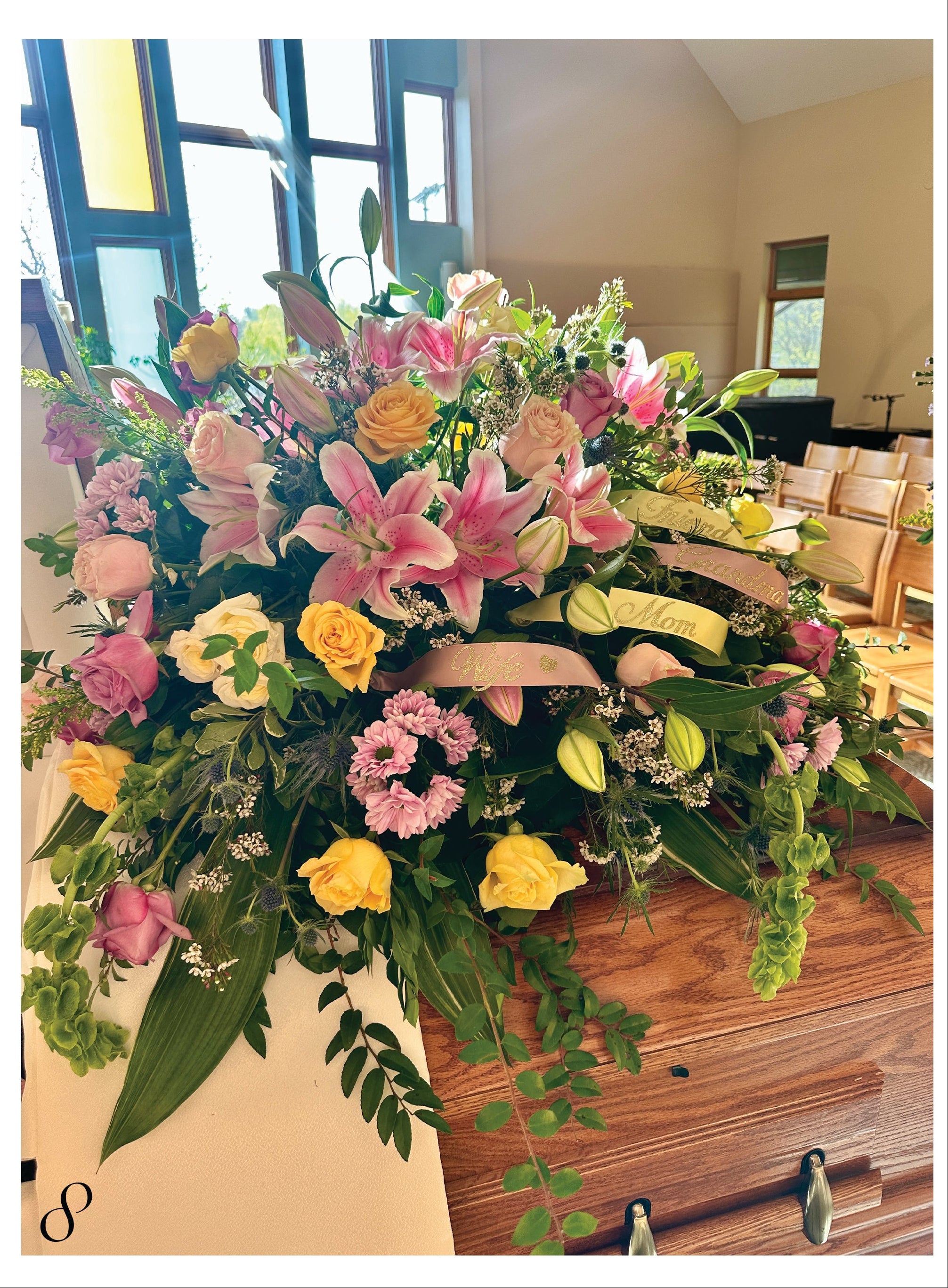 Colorful floral arrangement atop a wooden casket in a church setting with wooden pews.