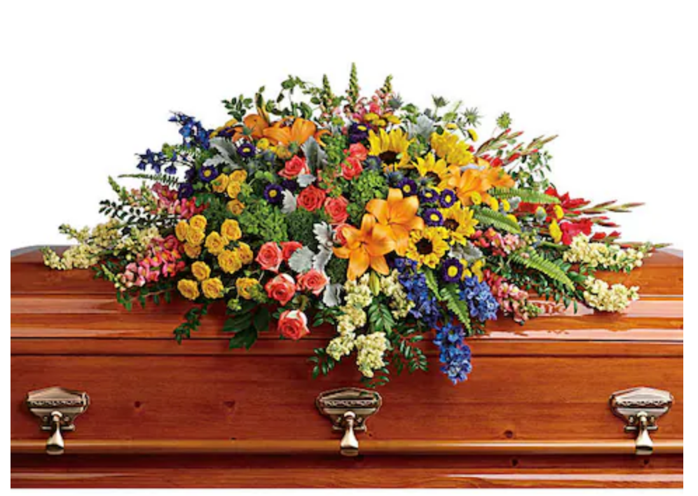 A colorful floral casket spray with a variety of flowers including hydrangea, roses, lilies, and sunflowers arranged on a wooden casket.