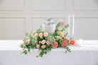 A floral centerpiece arrangement with a white urn, peach and pink roses, green foliage, white candles, and small white candles on a white table against a white paneled wall.
