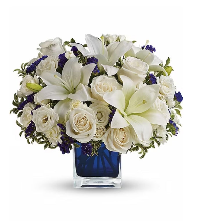 A bouquet of cream roses and white lilies arranged in a sapphire blue cube vase.