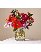 A brightly colored floral arrangement featuring a glass vase, with a variety of flowers including red and pink roses and other small blooms.